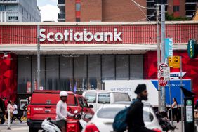 People and cars are seen on a street in front of a Scotiabank branch, with a red and white sign for the bank seen above the building.