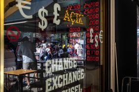 People are reflected in a window of a currency exchange that displays symbols for various currencies.