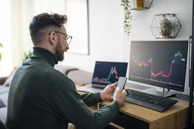 Person looking at a smartphone with stock graphs on two computer screens on his desk