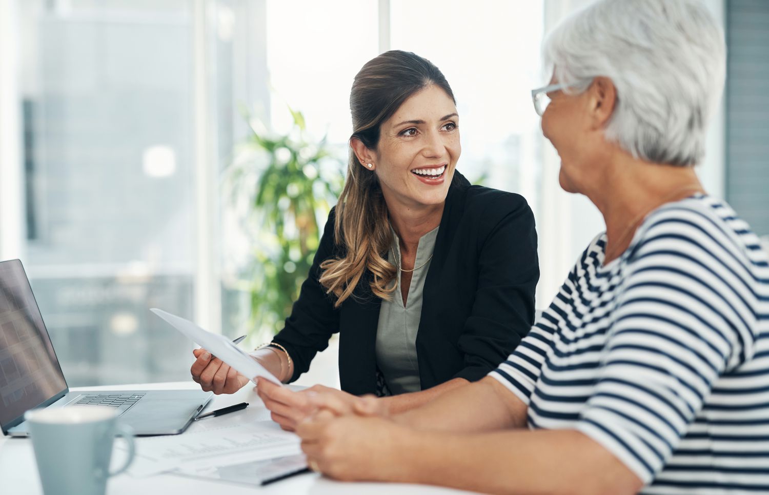 Young professional woman and an older woman sit in an office environment with a document and laptop.