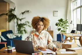 A young millennial woman sitting at her desk, with green plants in the background.
