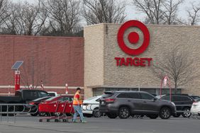 A Target worker pulls a group of shopping carts through a parking lot, with several cars and a Target store visible in the background.