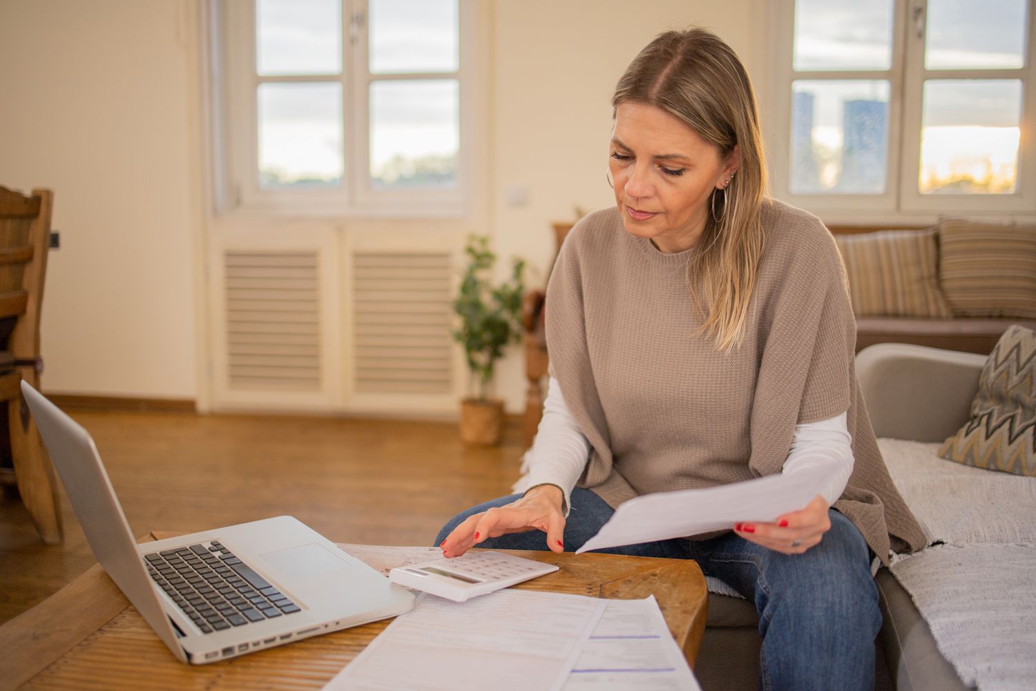 Woman in her 40s or 50s sitting on her sofa and looking at financial documents, a laptop, and a calculator