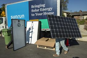 Two employees of Sunrun carry solar panels to be installed on the roof of a home, walking past a truck with a Sunrun logo.