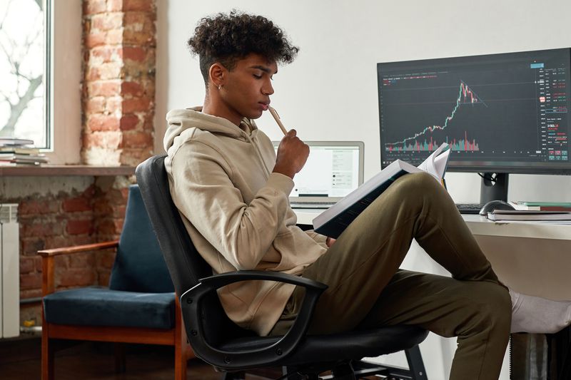 Young male sitting in front of monitors with financial data, looking at a notepad on his lap.