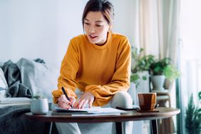 A woman in a bright setting writing on paper at a table with coffee and plants visible in the background