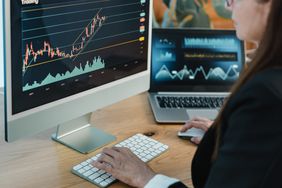 A woman types on a keyboard while sitting in front of a computer screen and laptop, both displaying financial charts. 