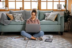 A woman sits on the floor in front of a green couch; she types on a laptop with papers, a tablet, and a mug on the floor around her
