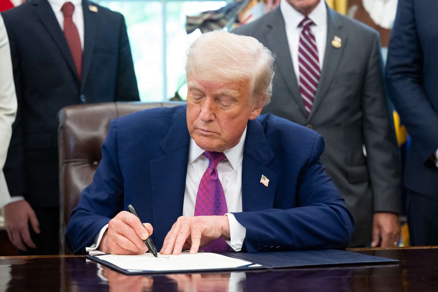 Donald Trump sitting at a desk, signing a document, with others standing in the background