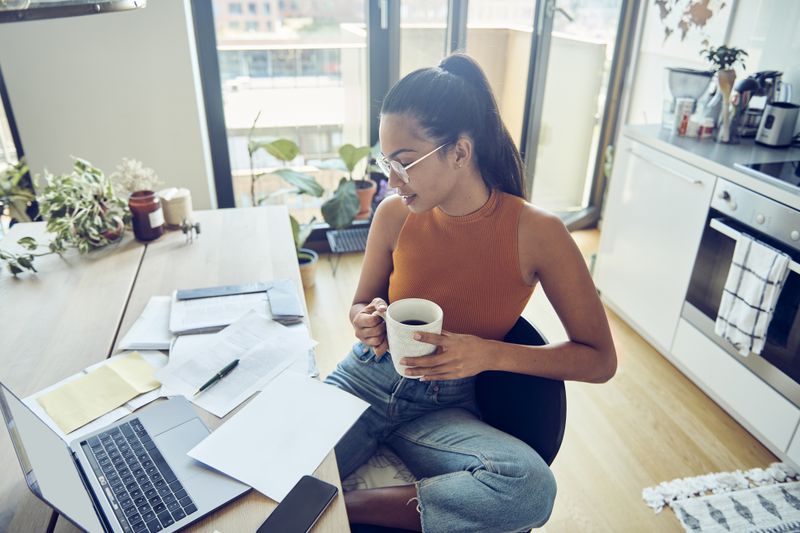 Woman sitting in a home office looking at papers on a desk with a laptop.