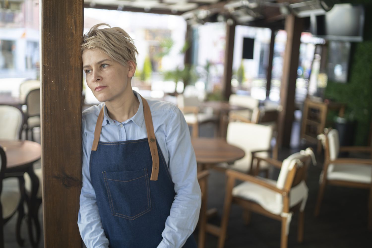 A worried restaurant server leans against a post in an empty restaurant worrying about their low income.