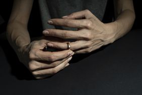 A dark background surrounding a woman's hands close up with one hand pulling off a wedding ring. 