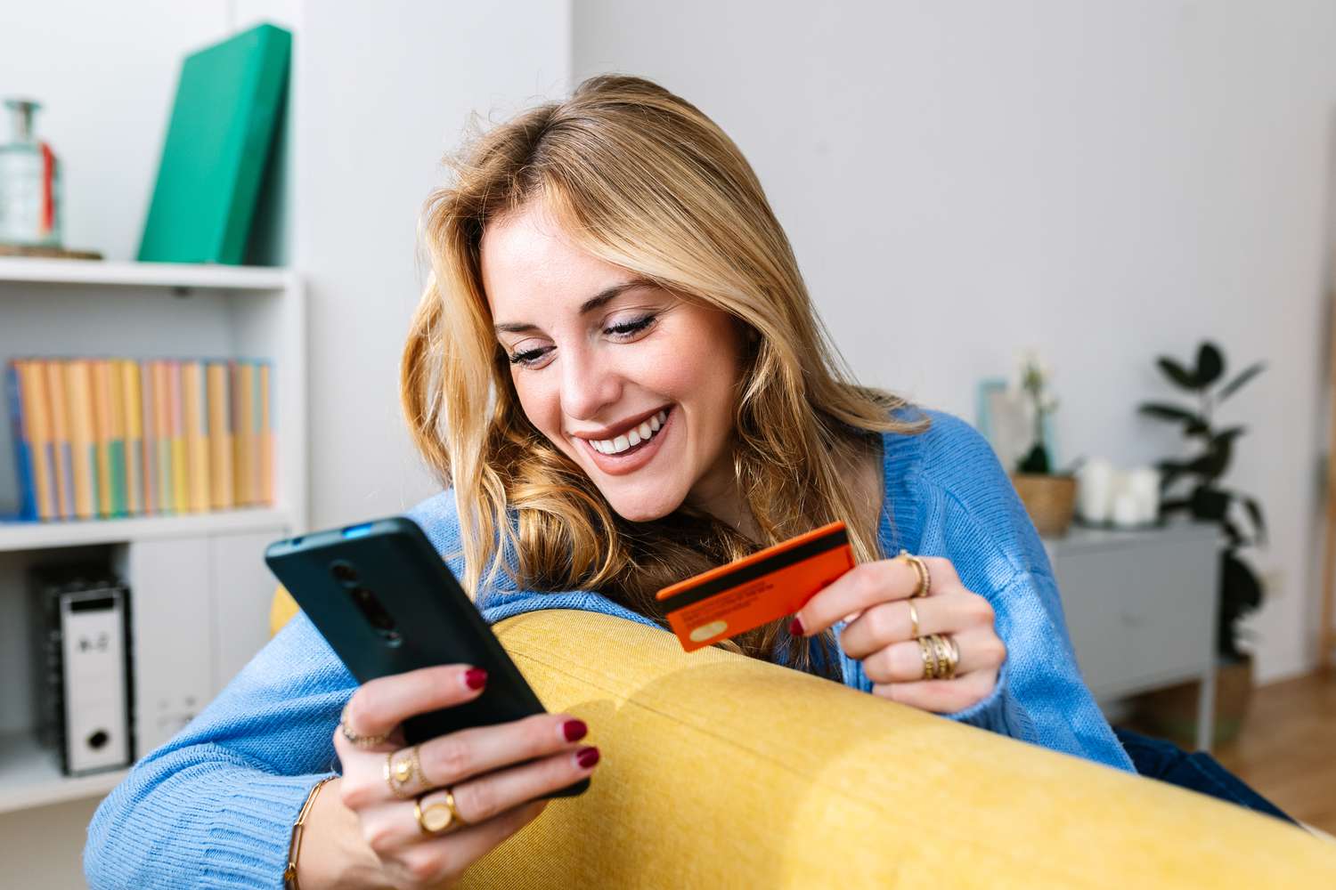 Similing young woman holding a phone and a credit card while sitting on a yellow couch. 
