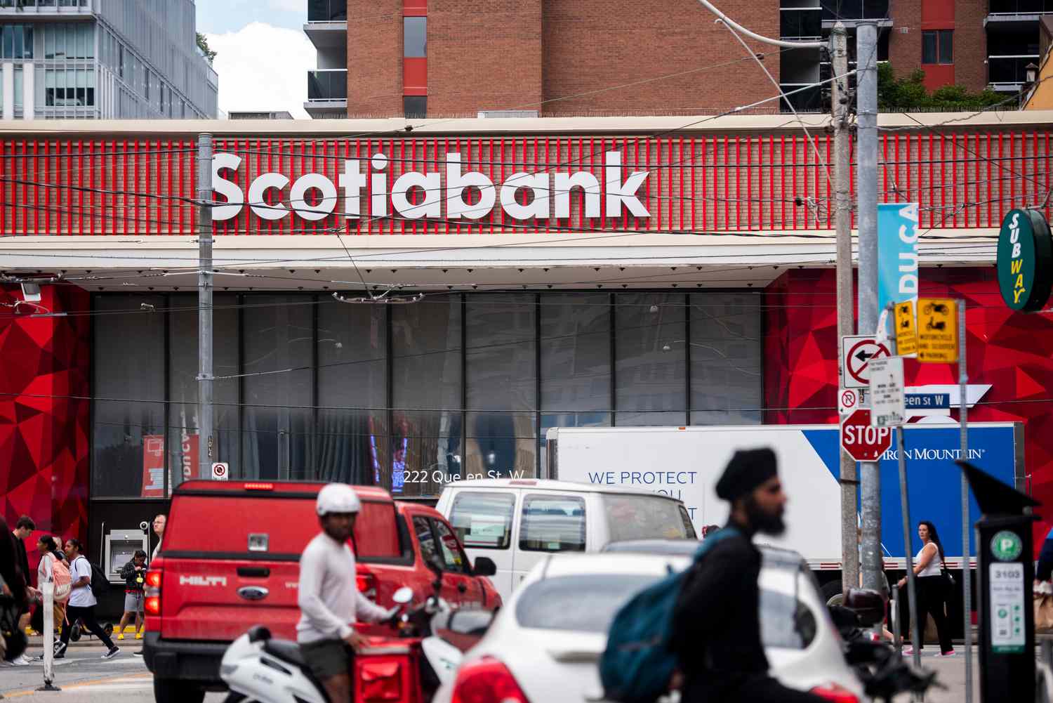 People and cars are seen on a street in front of a Scotiabank branch, with a red and white sign for the bank seen above the building.