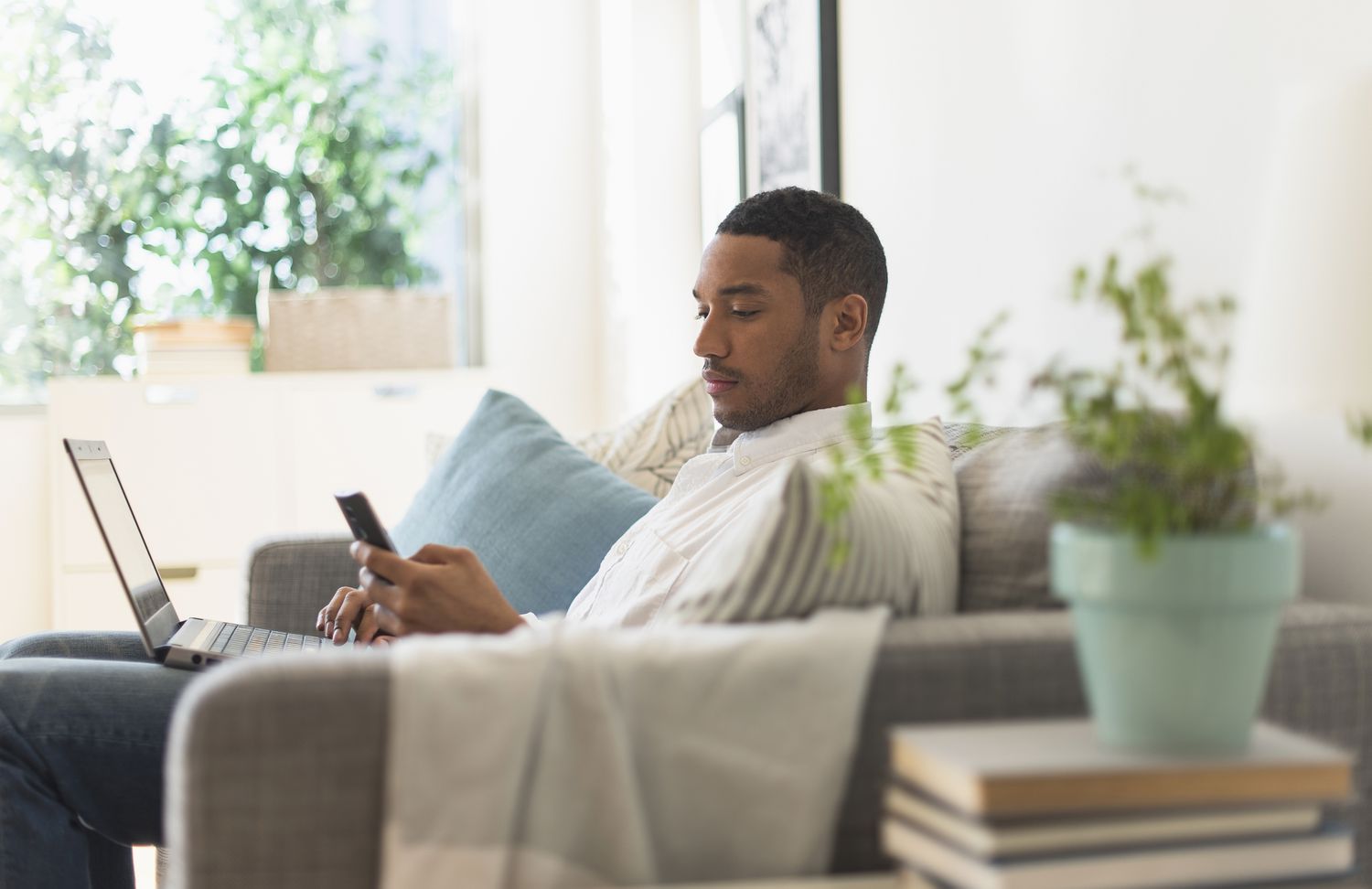 A man sits on a couch with a laptop looking at his smartphone