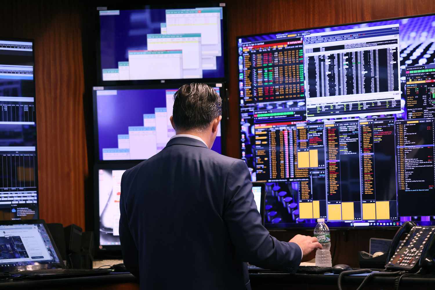 Man standing in front of a bank of monitors displaying market data.