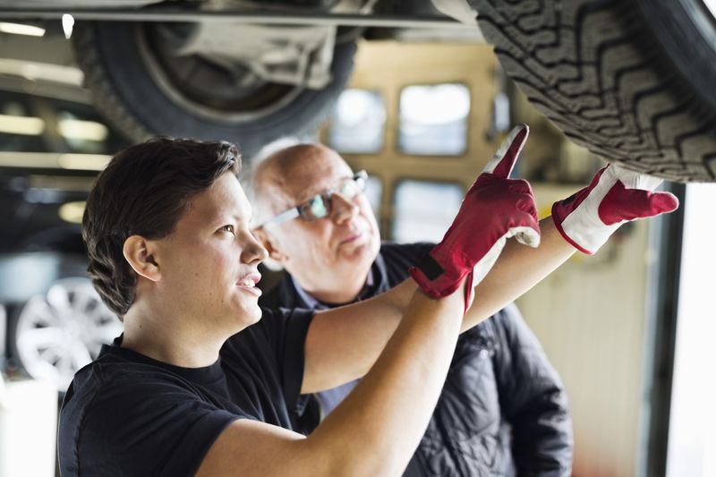 Young mechanic explaining tire problems to customer below car at auto repair shop