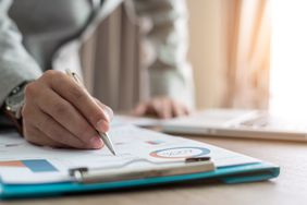 a person from the chest down writing on a clipboard with a pen. the clipboard shows graphs and charts