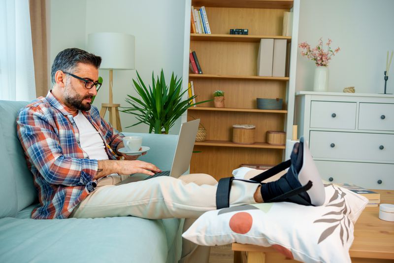 man working on laptop in home stretches broken foot in a cast on a pillow placed on the coffee table