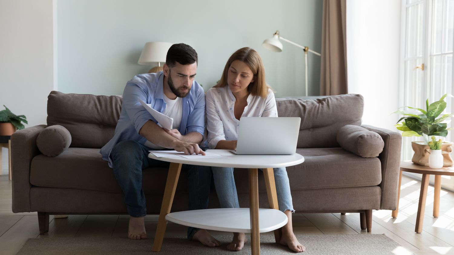 Millennial couple sitting on living room sofa and studying laptop and paperwork together