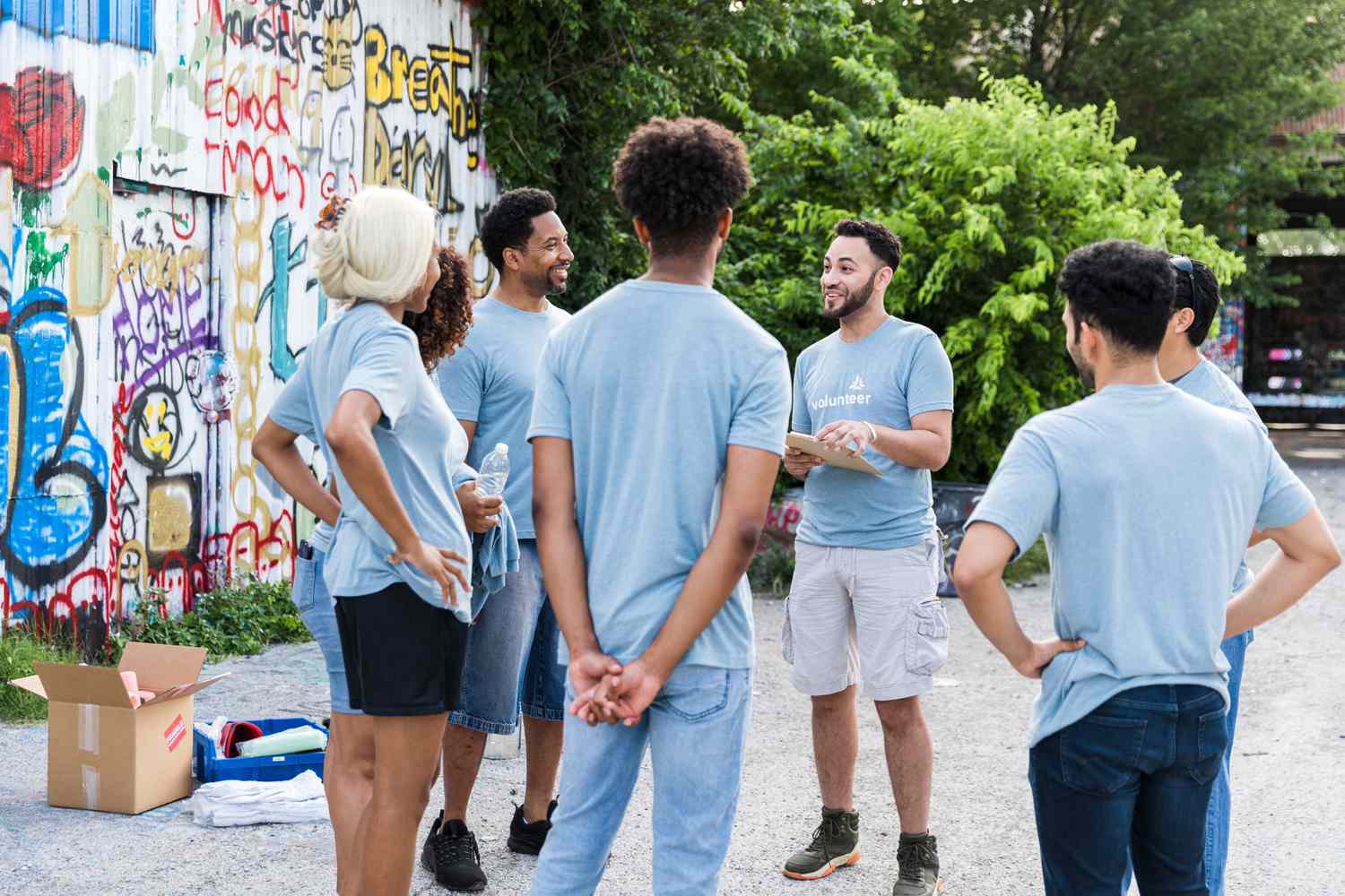 A project leader stands in an outdoor, graffitied empty lot and explains details to participants about a program funded by the government.