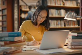 Young lady using a laptop to do research on the internet while sitting in a library surrounded by books