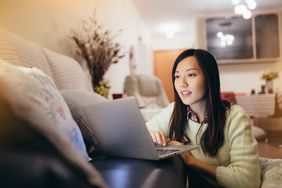 A woman wearing a white sweater sits on the ground facing a sofa looking at a laptop that is on the sofa