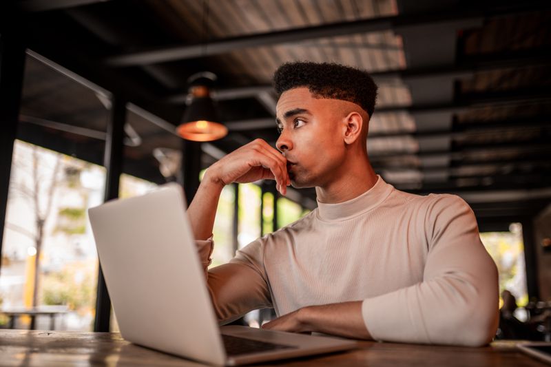 A young man sits at a laptop looking slightly worried.