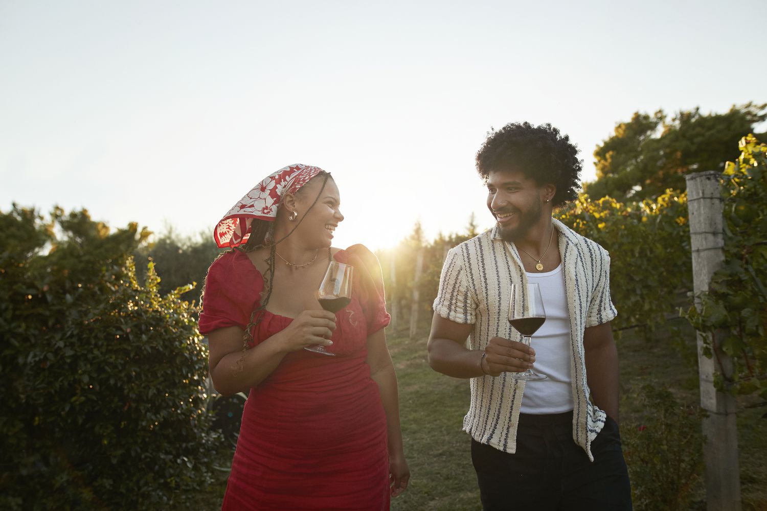 Two people holding wine glasses walking through a vineyard, smiling and enjoying the setting