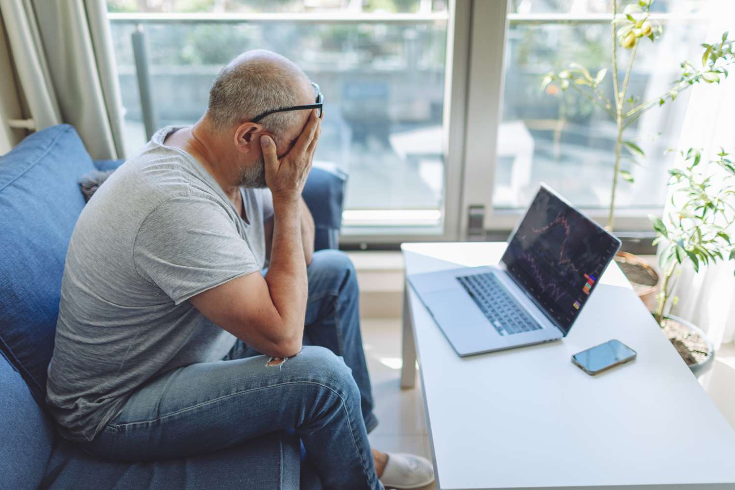 A despondent investor sits on their sofa covering their face with their hands after seeing a drastic decline in their market holdings on the laptop computer in front of them.