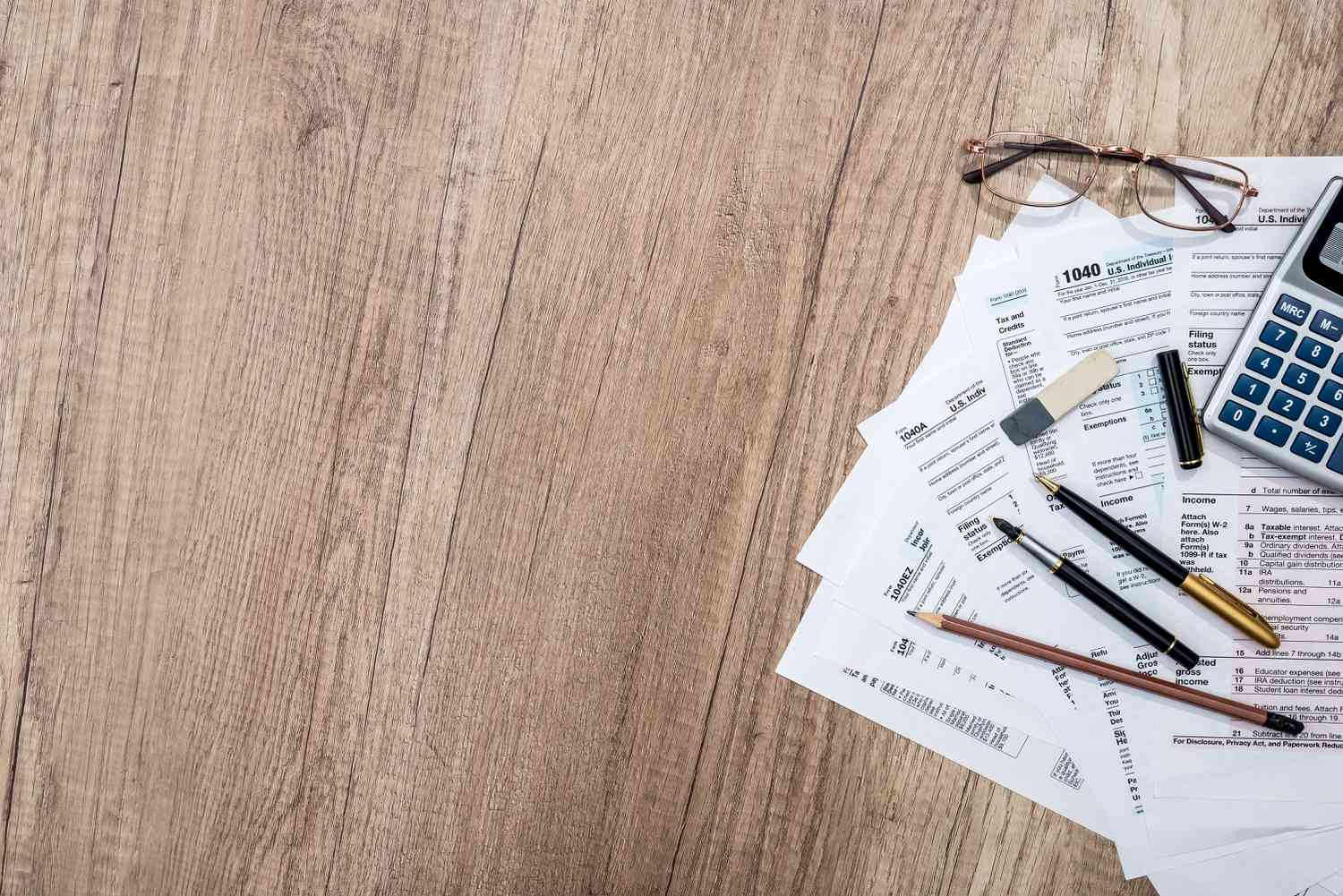 Tax forms on wooden table with notepad, pen, pencil, glasses, and calculator