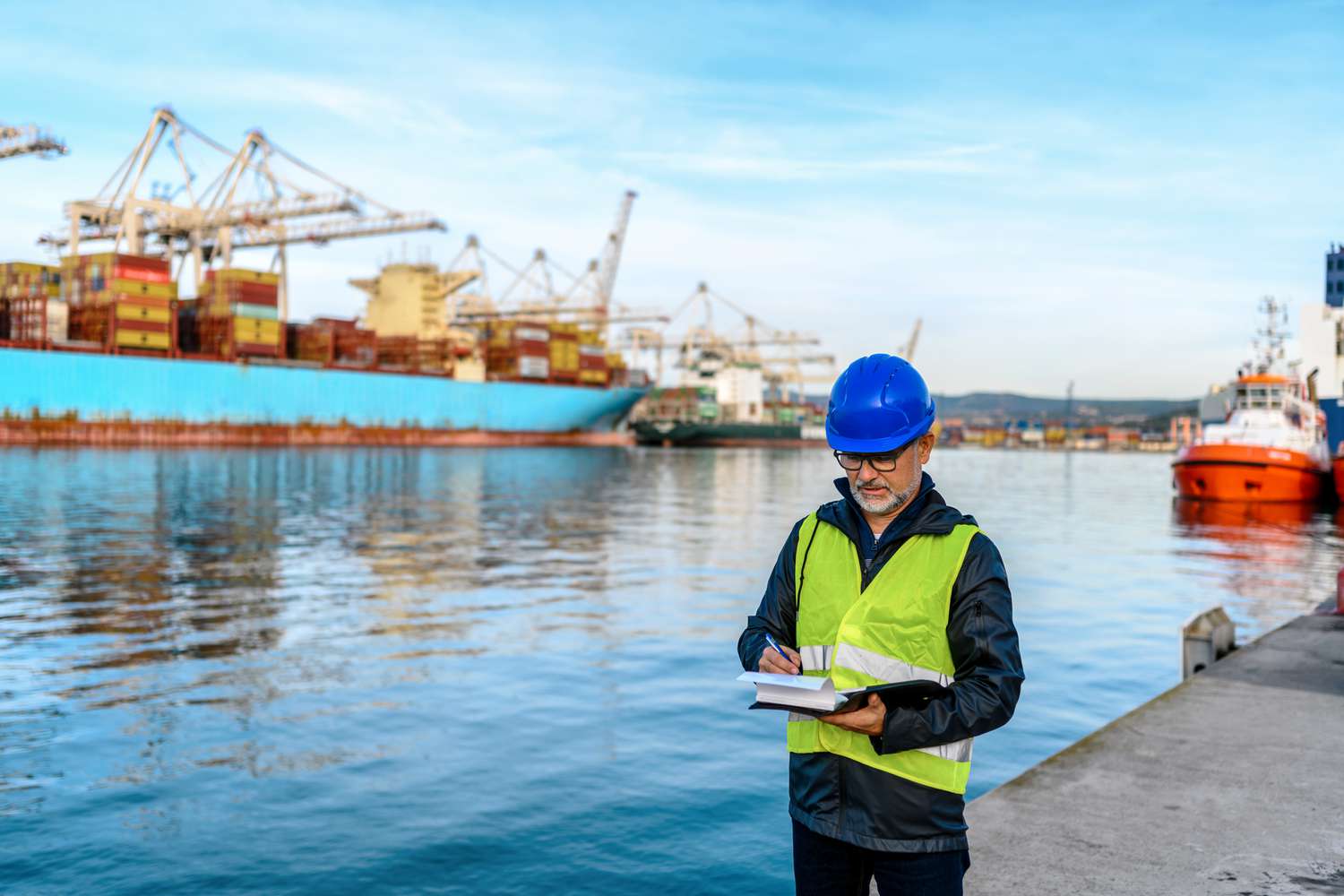 A consignee wears a hardhat and safety vest as they stand at an import terminal dock and reviews paperwork for an incoming cargo shipment.