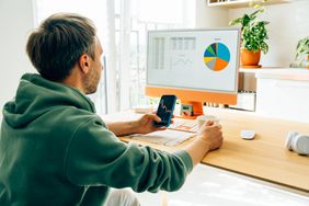 Man in home office looking at data on a monitor while holding a mobile phone.