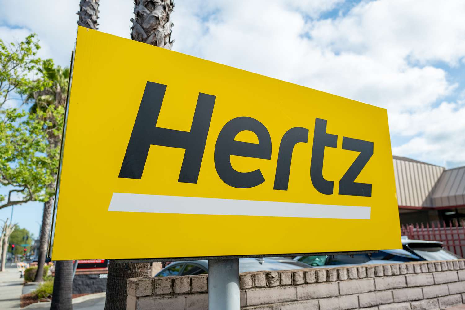 The yellow and black Hertz logo is seen on a sign outside a parking lot.
