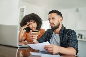 A man and woman sit at a table with a laptop, looking at a credit card.