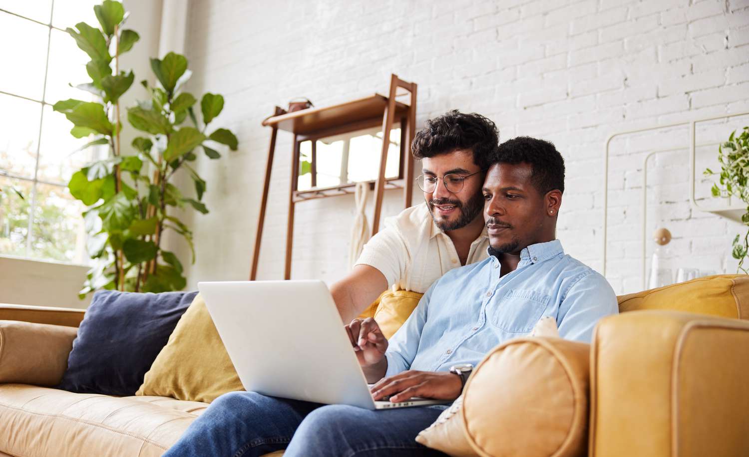A man sits on a couch with a laptop while another man stands behind him looking over his shoulder and pointing at the laptop