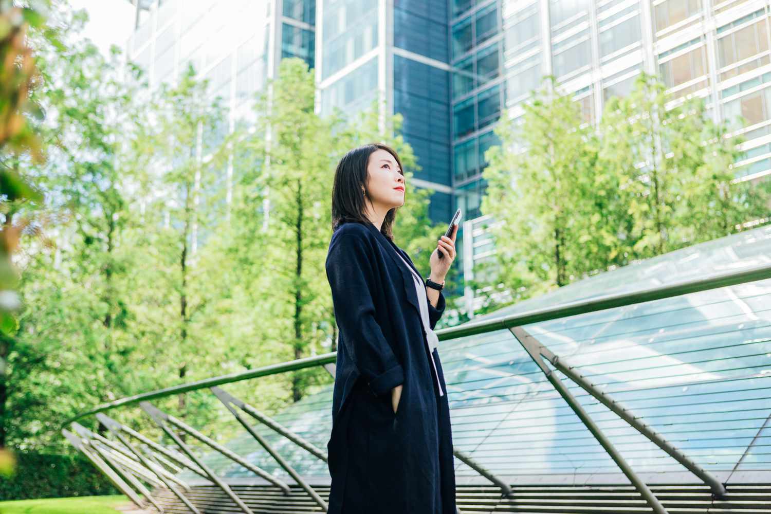 Young asian woman holding her smartphone, standing against corporate building surrounded by trees.