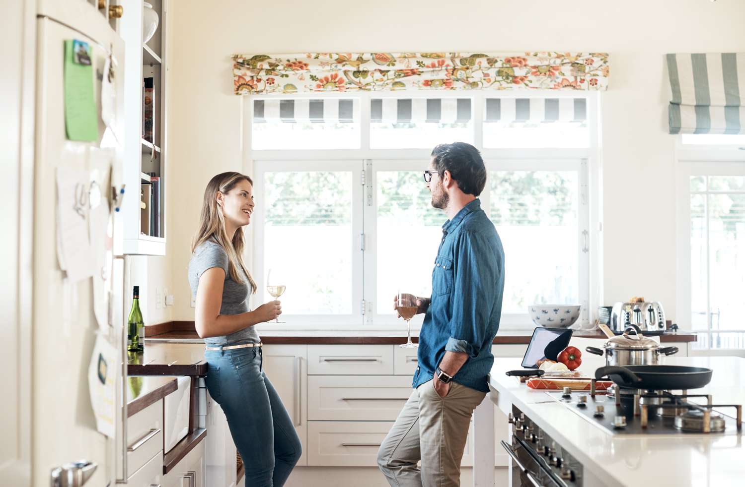A man and a woman stand in a kitchen talking while holding glasses of wine