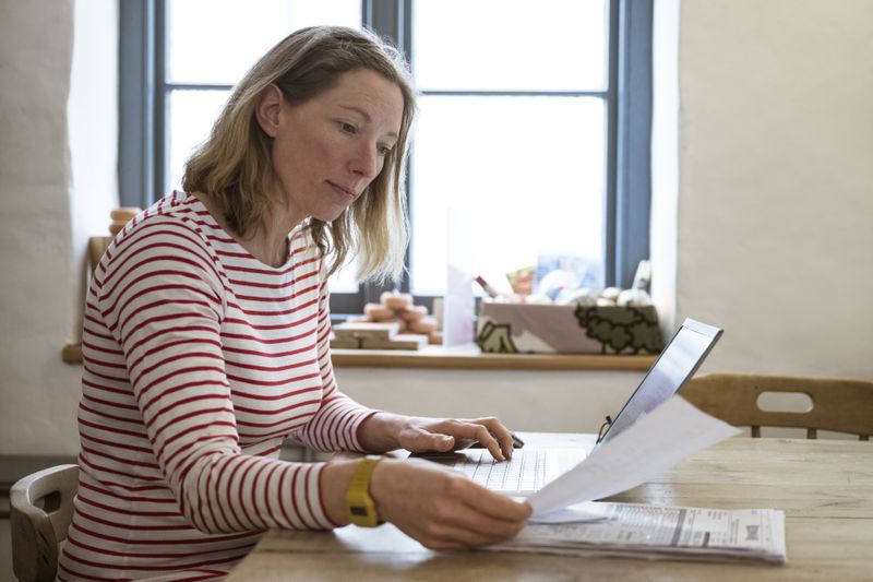 Woman in her 40s at her kitchen table, looking at financial documents and a laptop