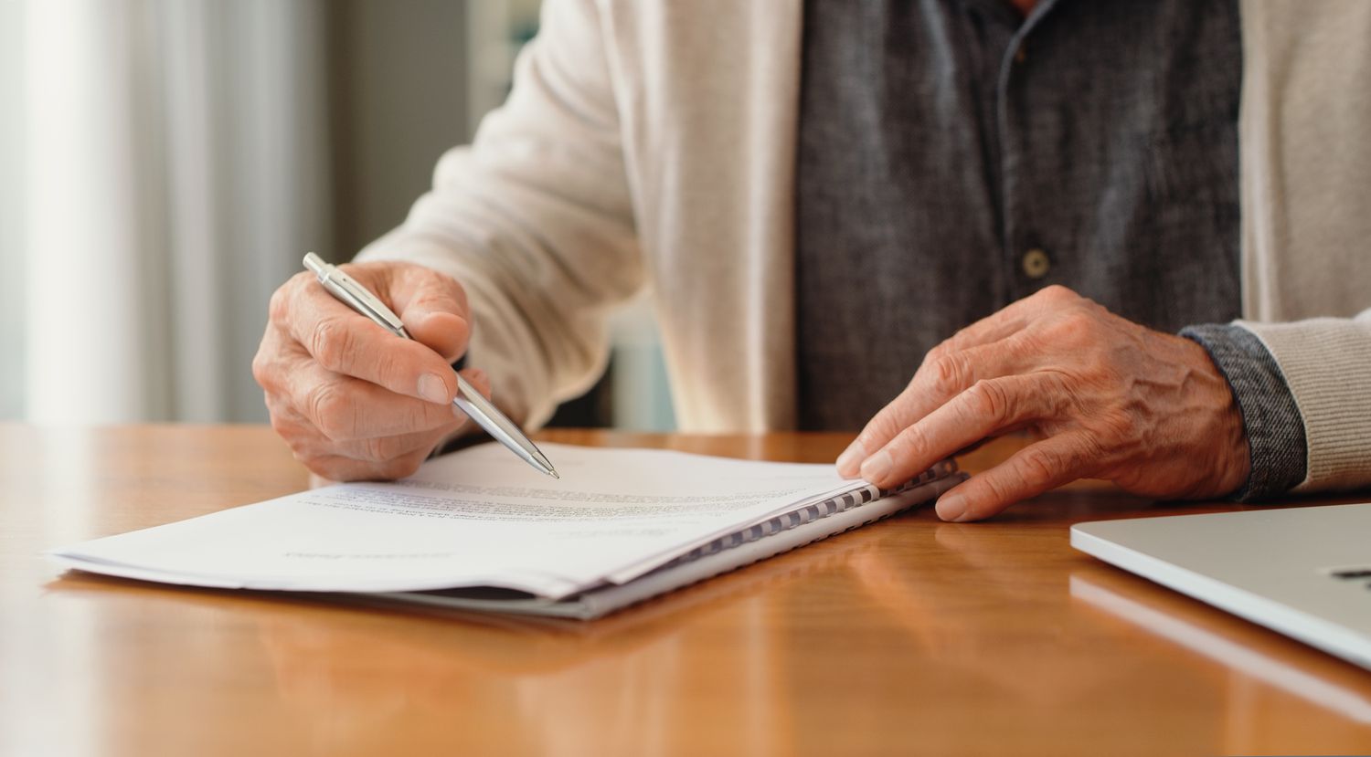 The hands of an older person as they read and sign their legal will while sitting at a table.