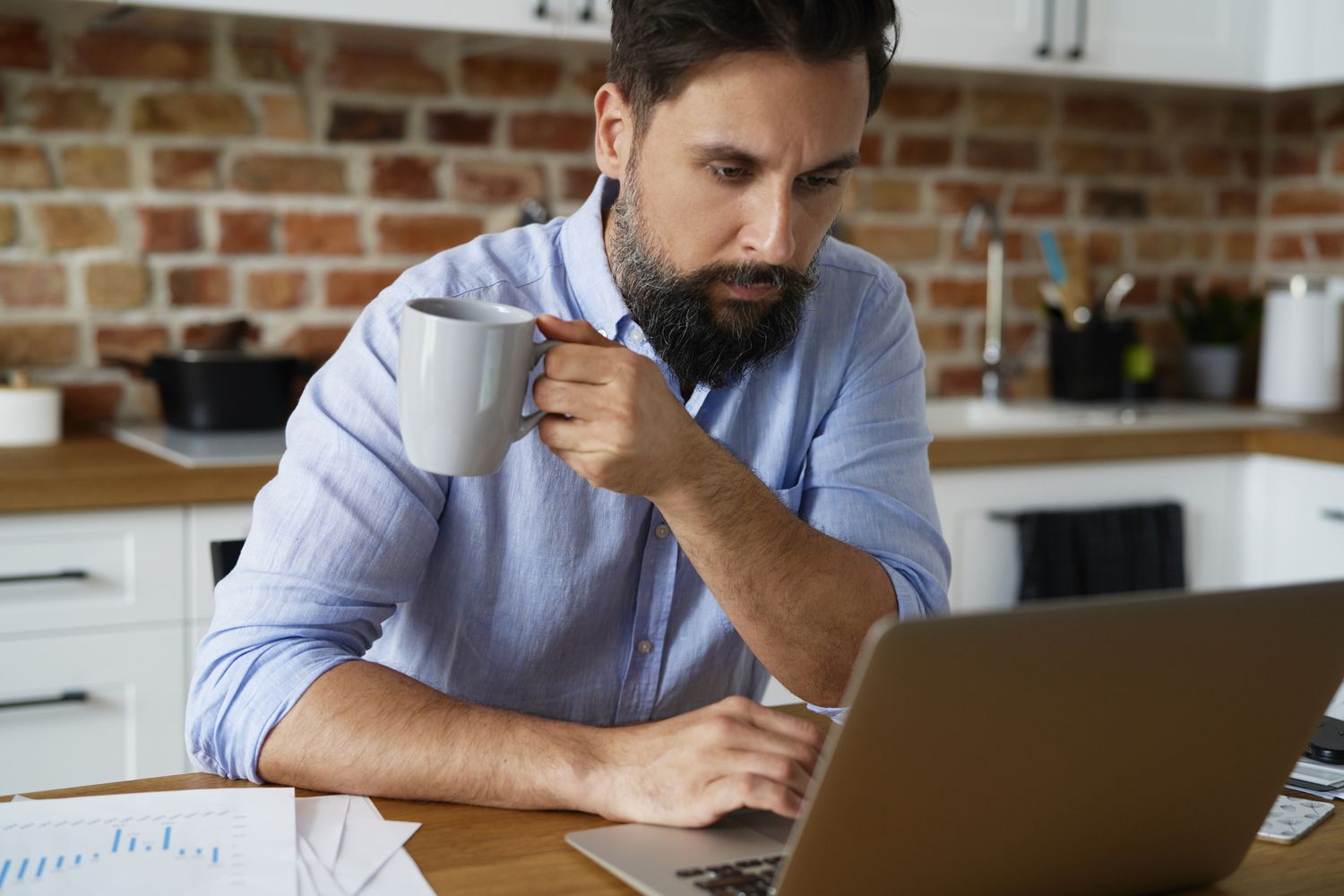 Man in his 30s or 40s sitting in his kitchen and looking seriously at mortgage rates on his laptop