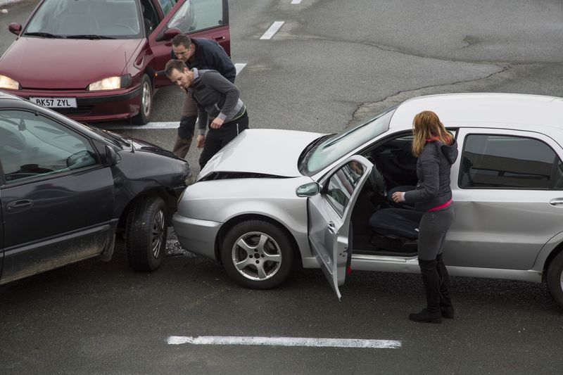 Two men looking at cars that have collided on a road, while a woman stands near one of them. 