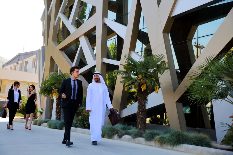 Two Middle Eastern business people walk and talk on a shaded sidewalk outside of a modern office building.