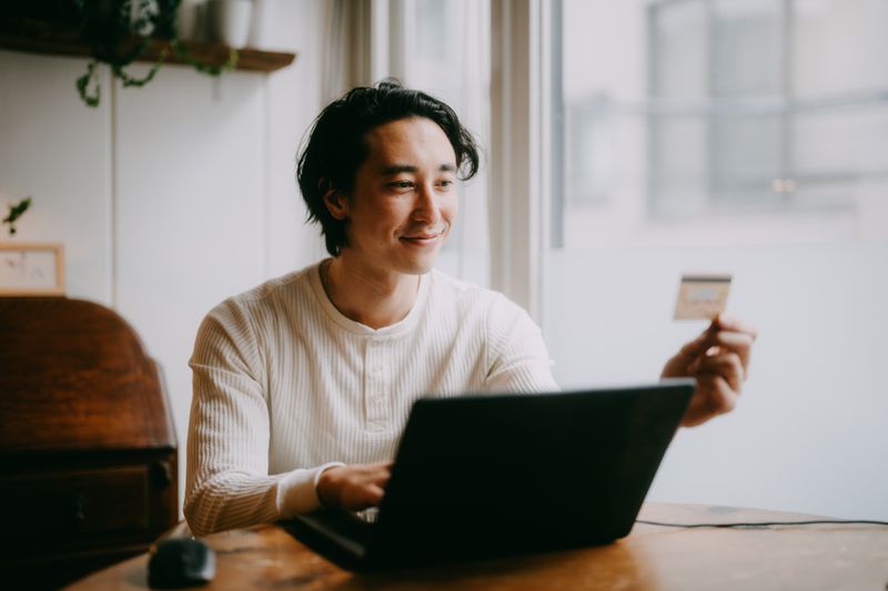 A man sits a table with a laptop, holding a credit card in his hand and smiling.