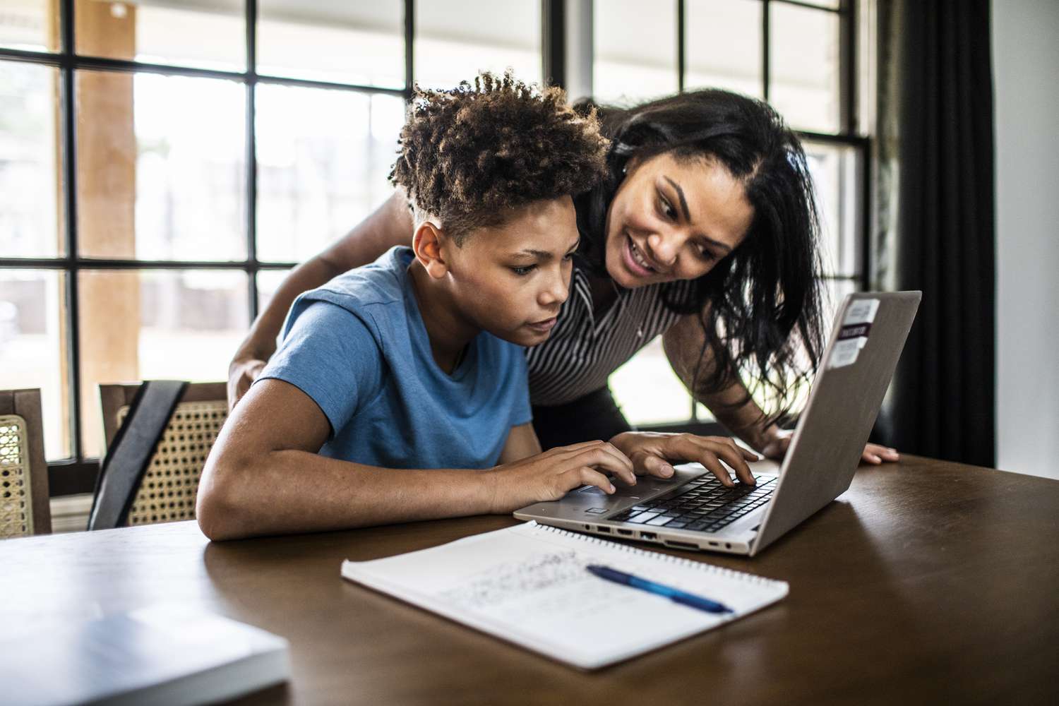 Adult woman helping younger man on laptop at a dining room table.