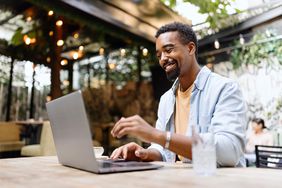 A happy man sits in a cafe using a laptop with a big smile on his face.