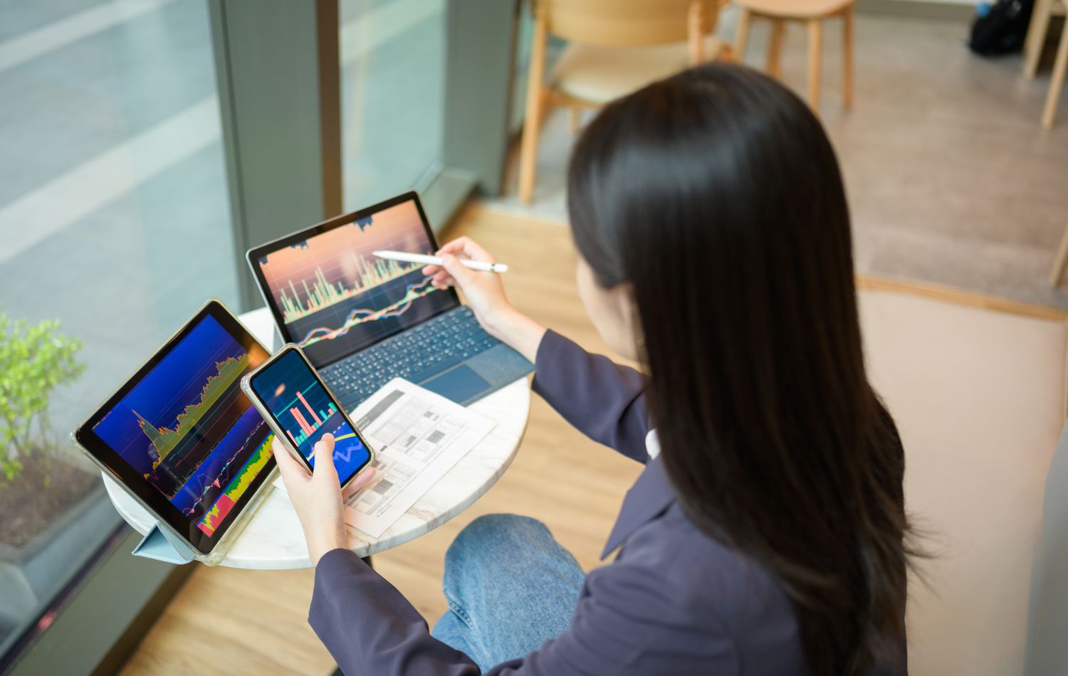 An investor sits at a table and uses a cellphone to enter a sell order as they track the price of the security on a computer screen.