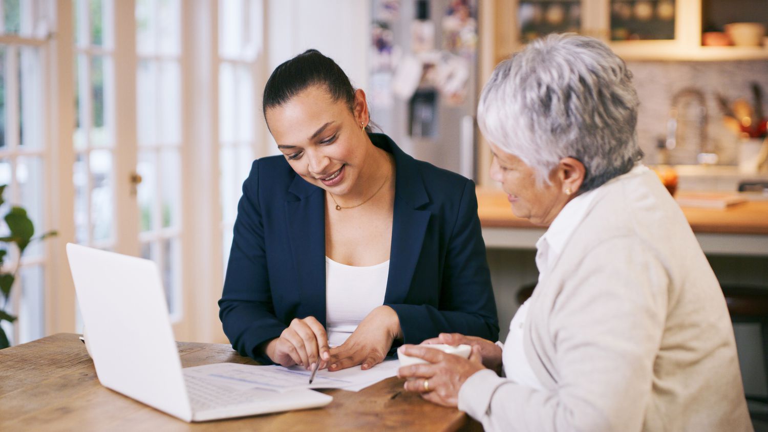 An older woman sitting at a kitchen table with a younger woman. There is a laptop and paperwork on the table that they are both looking at. 