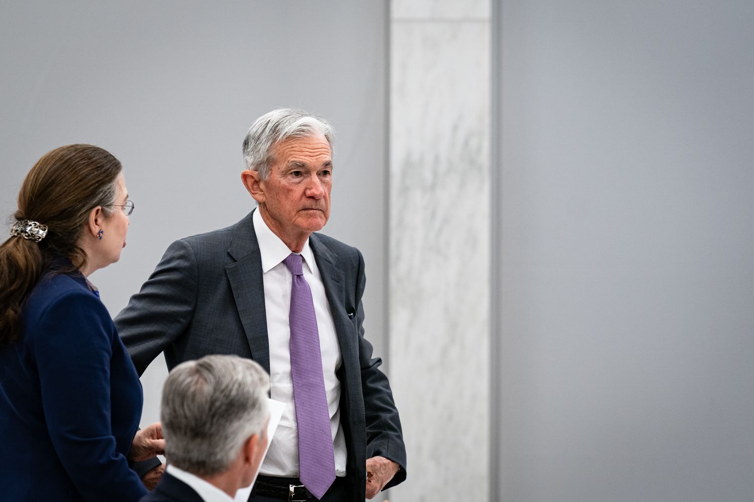 Jerome Powell standing while being addressed by a woman, another person seated in the foreground