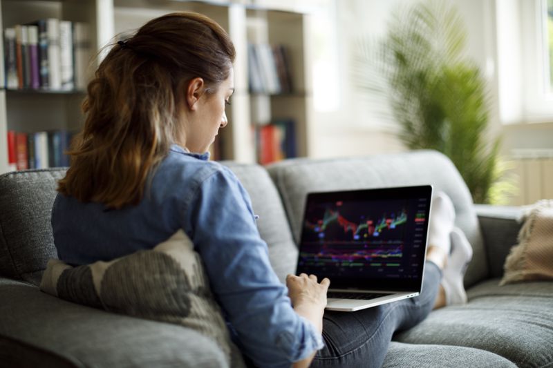 A woman with her back to the camera sits on a couch and looks at a laptop screen depicting stock charts.